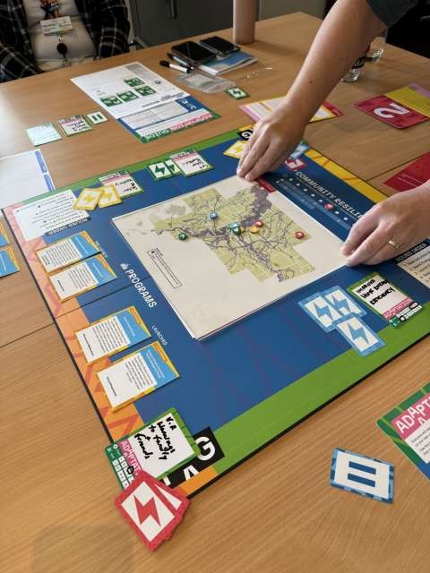 A close up of people playing the The Adaptation Game, showing the board game, cards and game pieces. 